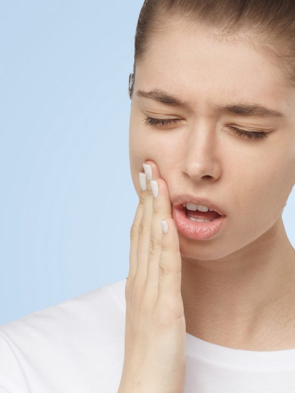 Young European woman isolated on blue background suffering from severe toothache, feeling pain so strong that she is pressing fingers to cheek to calm it down, looking desperate