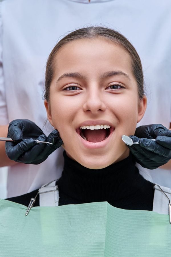 Portrait of young teenage girl in dental chair with hands of doctor with tools. Female teenager smiling with teeth looking at camera in dentist office. Adolescence hygiene treatment dental health care